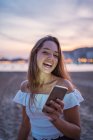 Heureuse jeune femme souriante et naviguant sur les médias sociaux sur smartphone tout en passant du temps sur la plage de sable fin en soirée — Photo de stock