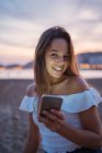 Feliz jovem mulher sorrindo e navegando mídias sociais no smartphone enquanto passa o tempo na praia de areia à noite — Fotografia de Stock