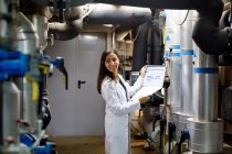 Seitenansicht einer jungen Frau im Laborkittel, die lächelt und wegschaut, während sie Daten im Journal während der Sicherheitsinspektion des Rohrleitungssystems in Industrieanlagen überprüft — Stockfoto
