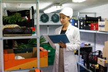 Donna in camice da laboratorio e cappello che prende appunti mentre controlla il cibo all'interno della cella frigorifera nel caffè — Foto stock