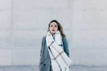 Giovane donna in cappotto elegante e sciarpa calda in piedi contro muro di cemento edificio e guardando in macchina fotografica sul tempo ventoso — Foto stock