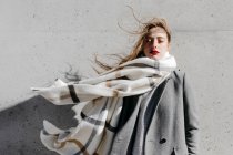 Jovem mulher em casaco elegante e cachecol quente de pé com os olhos fechados contra a parede do edifício de concreto em tempo ventoso — Fotografia de Stock