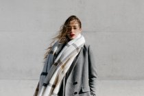 Молодая женщина в стильном пальто и теплом шарфе стоит с закрытыми глазами у бетонной стены здания в ветреную погоду — стоковое фото