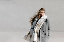Young woman in stylish coat and warm scarf standing with eyes closed against concrete building wall on windy weather — Stock Photo