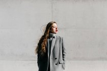 Jovem mulher de casaco cinza elegante de pé com os olhos fechados contra a parede do edifício de concreto em tempo ventoso — Fotografia de Stock