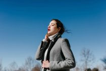 Боковий вид молодої жінки з закритими очима і в стильному сірому теплому пальто стояв проти чисте синє небо на вітряну погоду — стокове фото