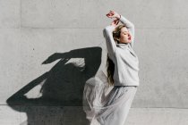 Вид збоку молодої жіночої моделі в стильному светрі і спідниці з закритими очима і піднятими руками проти сірої стіни на міській вулиці — стокове фото