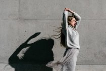 Side view of young female model in stylish sweater and skirt with eyes closed and raised arms against gray wall on city street — Stock Photo