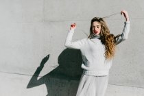 Молодая модель в стильном свитере и юбке с закрытыми глазами касающимися волос на серой стене на городской улице — стоковое фото