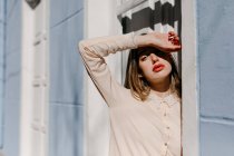 Молодая леди в элегантной блузке закрывает глаза от солнечного света, стоя рядом с белой дверью синего здания на улице — стоковое фото