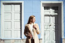 Giovane signora in camicetta elegante guardando altrove mentre in piedi vicino alla porta bianca di edificio blu sulla strada — Foto stock