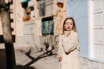 Young lady in elegant blouse with closed eyes standing near building on street in blurred background — Stock Photo