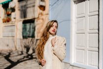 Молодая леди в элегантной блузке смотрит в сторону, стоя рядом со зданием на улице в размытом фоне — стоковое фото