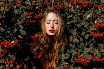 Mujer joven con los ojos cerrados de pie en medio de ramas verdes con bayas rojas en el día soleado en el jardín - foto de stock