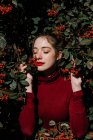 Молода жінка з закритими очима стоїть серед зелених гілок з червоними ягодами в сонячний день в саду — стокове фото
