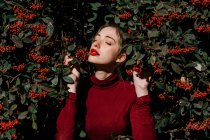 Молода жінка з закритими очима стоїть серед зелених гілок з червоними ягодами в сонячний день в саду — стокове фото
