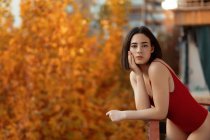 Hinreißende junge Frau in rotem Badeanzug lehnt an Geländer und blickt in die Kamera mit dem saisonalen herbstlichen verschwommenen Hintergrund — Stockfoto