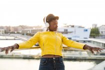 Zufriedene Afroamerikanerin in trendiger Jacke hört Musik über Kopfhörer, während sie sich auf gläsernen Balkon lehnt — Stockfoto