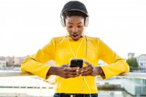 Afrikanisch-amerikanische Frau in stylischer heller Jacke benutzt Handy und hört Musik über Kopfhörer — Stockfoto