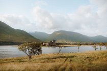 Danneggiato vecchio castello situato sulla costa del lago calmo contro colline erbose il giorno nuvoloso nella campagna britannica — Foto stock