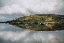 Picturesque landscape of mountain and cloudy sky reflected in tranquil water with sailboats in Glencoe on daytime — Stock Photo