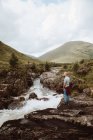 Вид сбоку на мужчину-туриста, стоящего на скалах над бурлящей рекой и созерцающего горный пейзаж в Гленко днем — стоковое фото