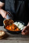 Crop person in apron standing by wooden table with skillet full of delicious butter chicken and rice — Stock Photo