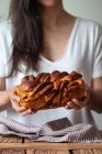Crop cuoco femminile in possesso di pane fresco torto o cannella babka sopra tavolo di legno con asciugamano a strisce su sfondo sfocato — Foto stock