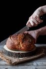Unrecognizable person using knife to cut loaf of fresh sourdough bread on piece of wood against black background — Stock Photo