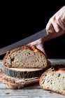 Pessoa irreconhecível usando faca para cortar pão de massa fresca em pedaço de madeira contra fundo preto — Fotografia de Stock
