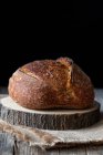 Loaf of fresh country sourdough bread placed on piece of wood on shabby table against black background — Stock Photo