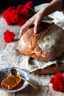 Unrecognizable person putting loaf of sourdough bread on rustic table near honeycomb and bouquet of red carnations — Stock Photo