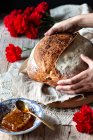 Unrecognizable person putting loaf of sourdough bread on rustic table near honeycomb and bouquet of red carnations — Stock Photo
