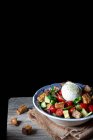 Ciotola con deliziosa insalata di panzanella posta su un panno su un tavolo di legno su sfondo nero — Foto stock