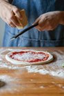Chico irreconocible en delantal moliendo queso fresco en masa con salsa de tomate mientras prepara pizza en la mesa - foto de stock