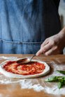Chef anonimo che spalma sugo di pomodoro fresco sulla pasta cruda mentre cucina la pizza in tavola — Foto stock