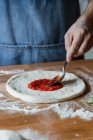 Anonymous chef smearing fresh tomato sauce on raw dough while cooking pizza on table — Stock Photo