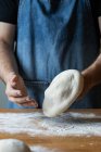 Неузнаваемый мужчина в фартуке сглаживает мягкое тесто за столом с мукой во время приготовления пиццы — стоковое фото