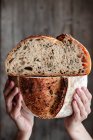 Personne méconnaissable montrant du pain frais coupé en deux avec des graines contre un mur en bois — Photo de stock
