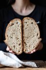 Personne méconnaissable montrant du pain frais coupé en deux avec des graines contre un mur en bois — Photo de stock