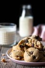 Assiette de biscuits savoureux avec des pépites de chocolat placées sur la table de bois près du lait flou et du tissu sur fond noir — Photo de stock