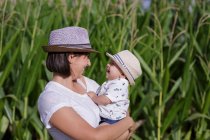 Vista lateral de la madre alegre elegante y el niño en la mano que tienen tiempo juntos en el campo agrícola verde - foto de stock