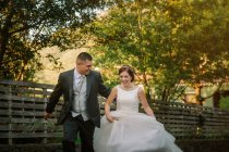 Елегантний веселий наречений тримає чарівну наречену і бігає разом зі сміхом по дорозі вздовж дерев'яного паркану в зеленому саду — стокове фото