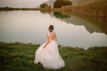 Vue arrière de la mariée tenant robe gracieuse avec dos ouvert et regardant calme lac de cristal reflétant ciel — Photo de stock