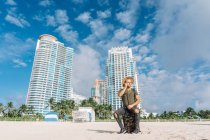 Touriste de sexe masculin assis sur sa valise à la plage, Immeubles et vue époustouflante sur le ciel — Photo de stock
