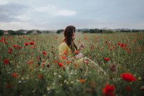 Pensive attractive red haired adult lady in yellow dress with red poppy in hair and red lips looking away while sitting alone in blurred amazing green meadow with red and white flowers against hills under gray cloudy sky — Stock Photo