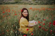 Красивая привлекательная рыжеволосая взрослая дама в желтом платье с рыжими волосами и рыжими губами, глядя через плечо на камеру, сидя одна в размытом удивительном зеленом меде с красными и белыми цветами на фоне холмов под облачным небом — стоковое фото