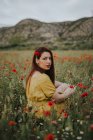 Pensive attractive red haired adult lady in yellow dress with red poppy in hair and red lips looking over shoulder at camera while sitting alone in blurred amazing green meadow with red and white flowers against hills under gray cloudy sky — Stock Photo