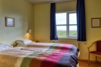 Dos cómodas camas aseadas con almohadas blancas y mantas de colores cálidos en la habitación acogedora luz con paredes amarillas contra la ventana con cortinas azules con vistas al paisaje rural - foto de stock