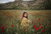 Pensive attractive red haired adult lady in yellow dress with red poppy in hair and red lips looking over shoulder at camera while sitting alone in blurred amazing green meadow with red and white flowers against hills under gray cloudy sky — Stock Photo